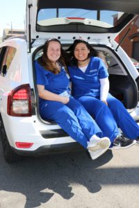 Two female students, wearing blue nursing scrubs, smile and pose while sitting out on the end of an open hatchback car