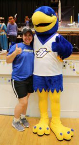 A female student wearing a blue shirt and black shorts hugs and poses with the blue and gold Spalding eagle mascot while both give thumbs up