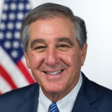 Head shot of Jerry Abramson, wearing coat and tie and smiling, with an American flag in background