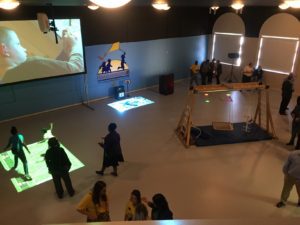 Overhead view of the Virtual Immersive Playground during its opening reception - a large open space, dimly lit, so as to show various video projections on the floors and walls; people mingling about