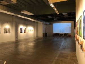 A view of an art gallery with paintings and drawings hung on the wall to the left, a display on a large screen on a center wall and several sculptures resembling human arms and hands on the right wall.