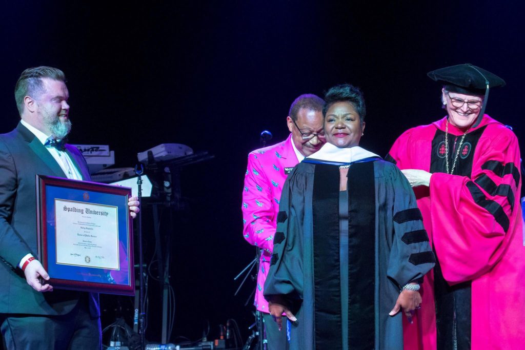 On stage, Sadiqa Reynolds, wearing black robe, is presented with an honorary degree from Spalding President Tori McClure, in a red robe, while Spalding Chief Advancement Officer, smiling and wearing a dark tuxedo, holds a framed Spalding diploma