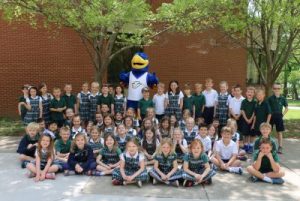 Dozens of smiling girls and boys in green and blue school uniforms pose outside under a tree with the big Spalding gold-and-blue eagle mascot
