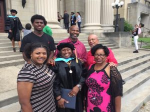 Cynthia Eddings-King, in black cap and gown, standing on steps, surrounded by smiling family