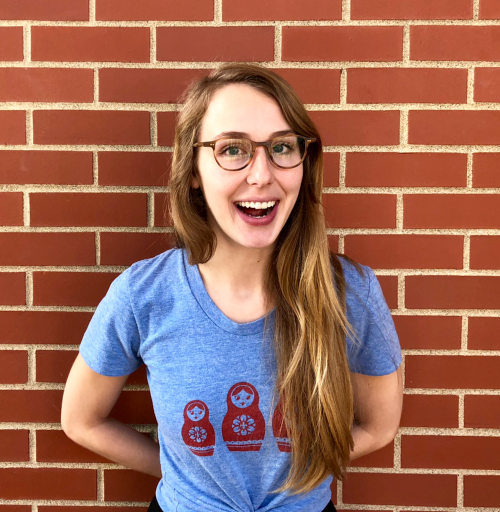 Anna Foshee, smiling and wearing glasses, standing in front of red brick wall