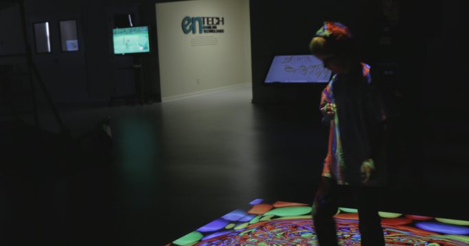 A young boy in a dark room walks on a bright, colorful floor that's illuminated with gesture-recognition video technology