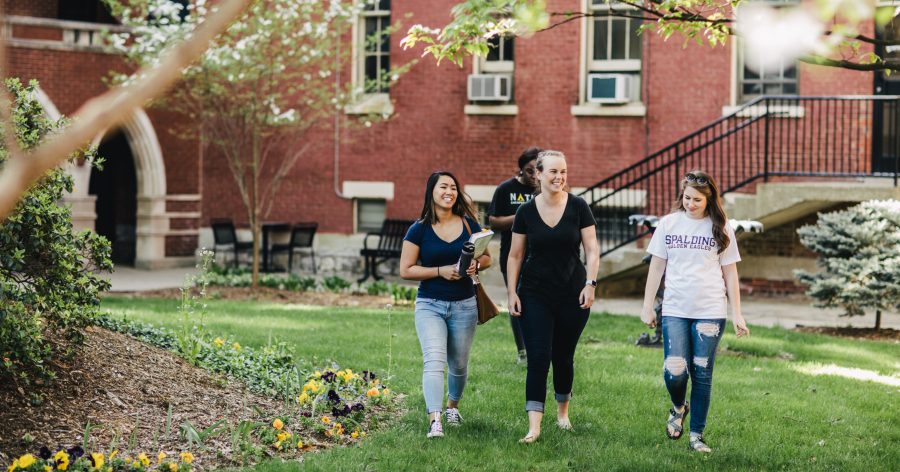 Three smiling female students walk across a campus lawn