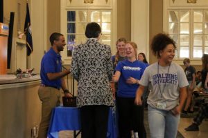 Smiling man Jeffrey Cross) and woman Spalding President Tori McClure) stand and distribute Spalding Mission coins to three female freshman college students