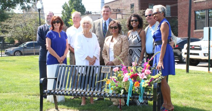 Men and women stand behind a black metal Trager Park bench, with bouquet set on top