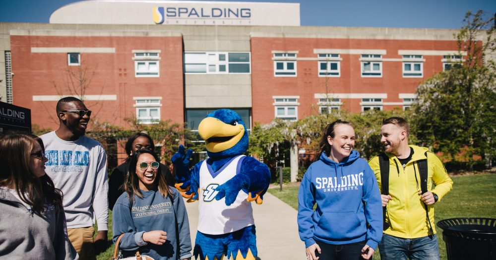 Six smiling college students and the blue Spalding Eagle mascot walk outside in front of a campus building that says "SPALDING"