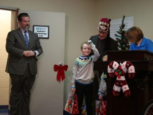 A young boy holding a gift bag smiles while standing next to a Christmas tree, Kosair Charities President Keith Inman, enTECH therapist Alison Amshoff and Spalding Advancement Chief Bert Griffin