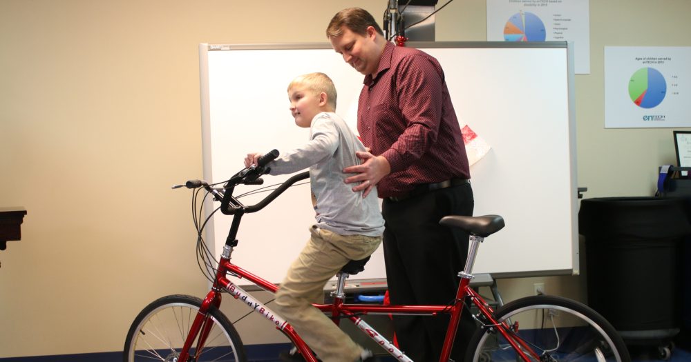 A father helps his young son onto a Buddy Bike tandem bicycle inside the enTECH lab