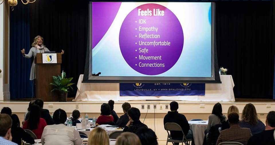 Western Middle School counselor Judith Wilson speaks to a crowd from a podium on stage as a PowerPoint presentation listing different emotions displays on a large screen next to her.
