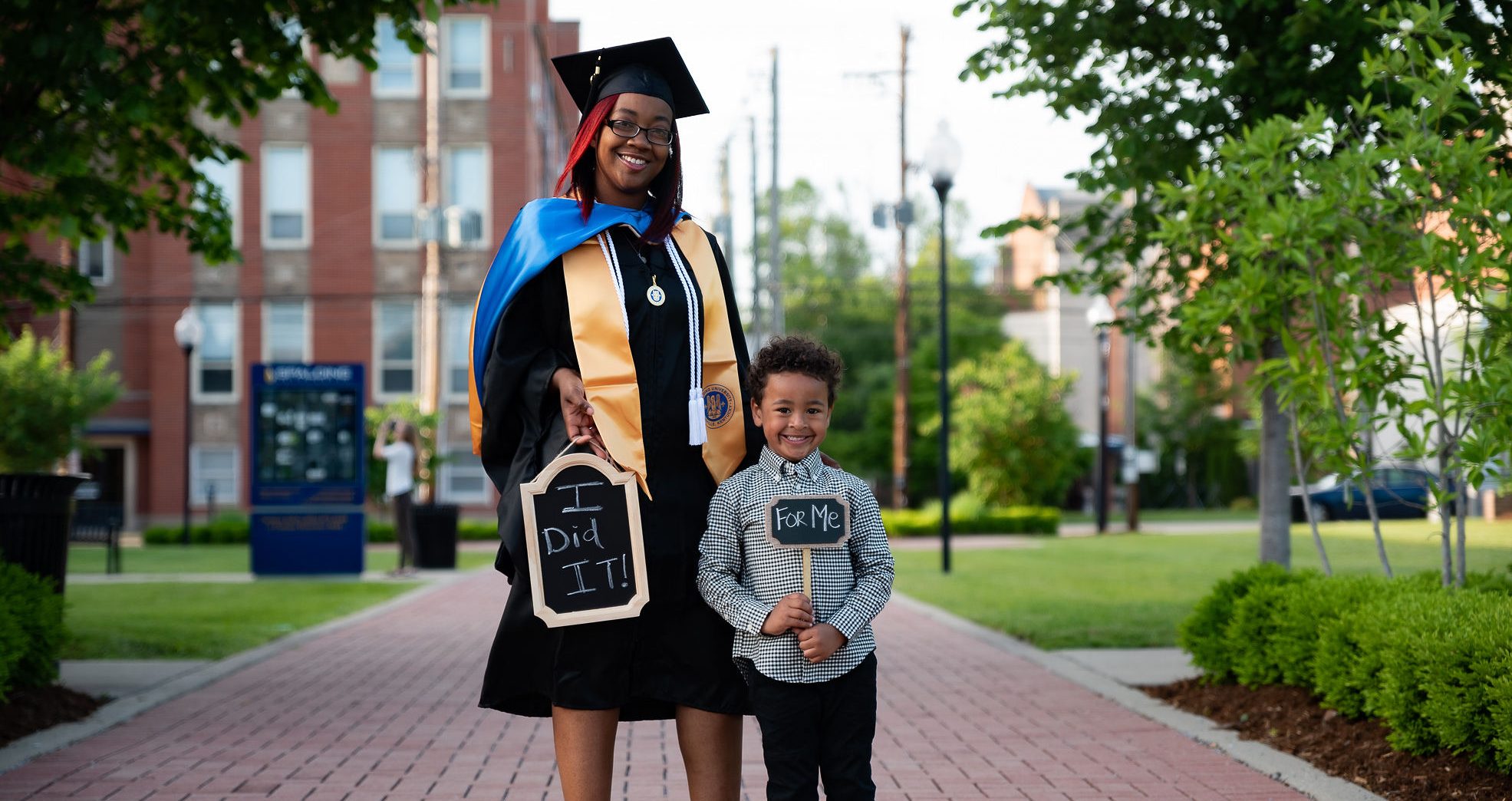 Spalding MSBC graduating student Kristin Spencer, wearing cap and gown, and her young son outside at Mother Catherine Square. Shes holding a sign, "I Did It!," and he has one that says, "For Me"