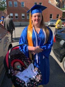 Spalding student Megan Faust, in blue cap and gown, holding baby, Madalyn, in a car seat