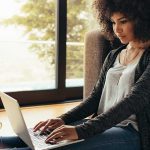 Online student sits in front of couch, works on laptop