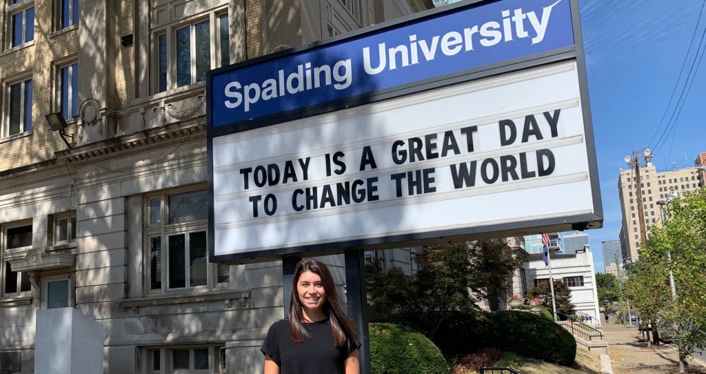 Student Miranda Wray standing outside under Spalding University sign that reads TODAY IS A GREAT DAY TO CHANGE THE WORLD