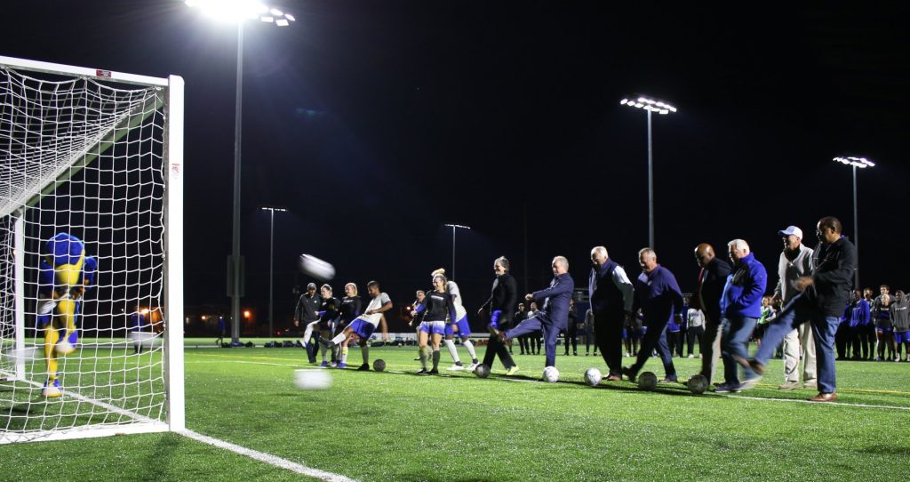 Row of people ceremoniously kick a soccer ball into a goal at the Spalding mascotwith a night sky behind them