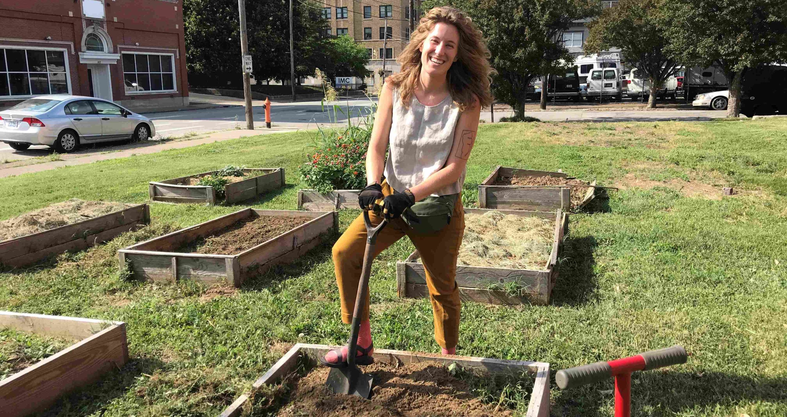 Spalding Librarian Leah Cover smiles and stands while holding a shovel in the ground of a raised bed in the Second Street Garden