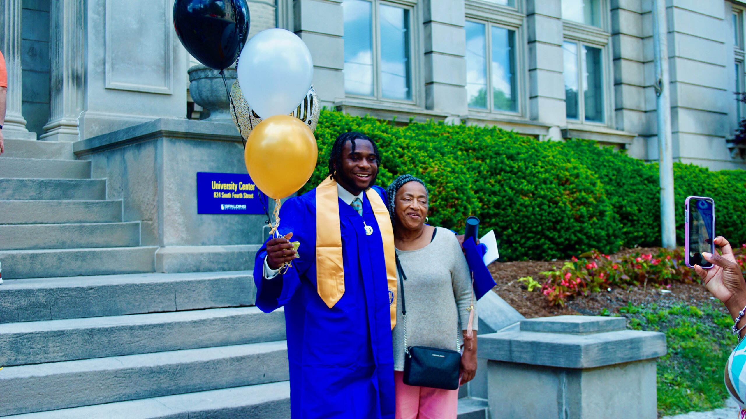 Spalding bachelor's graduate in cap and gown posing outside Columbia Gym with mom