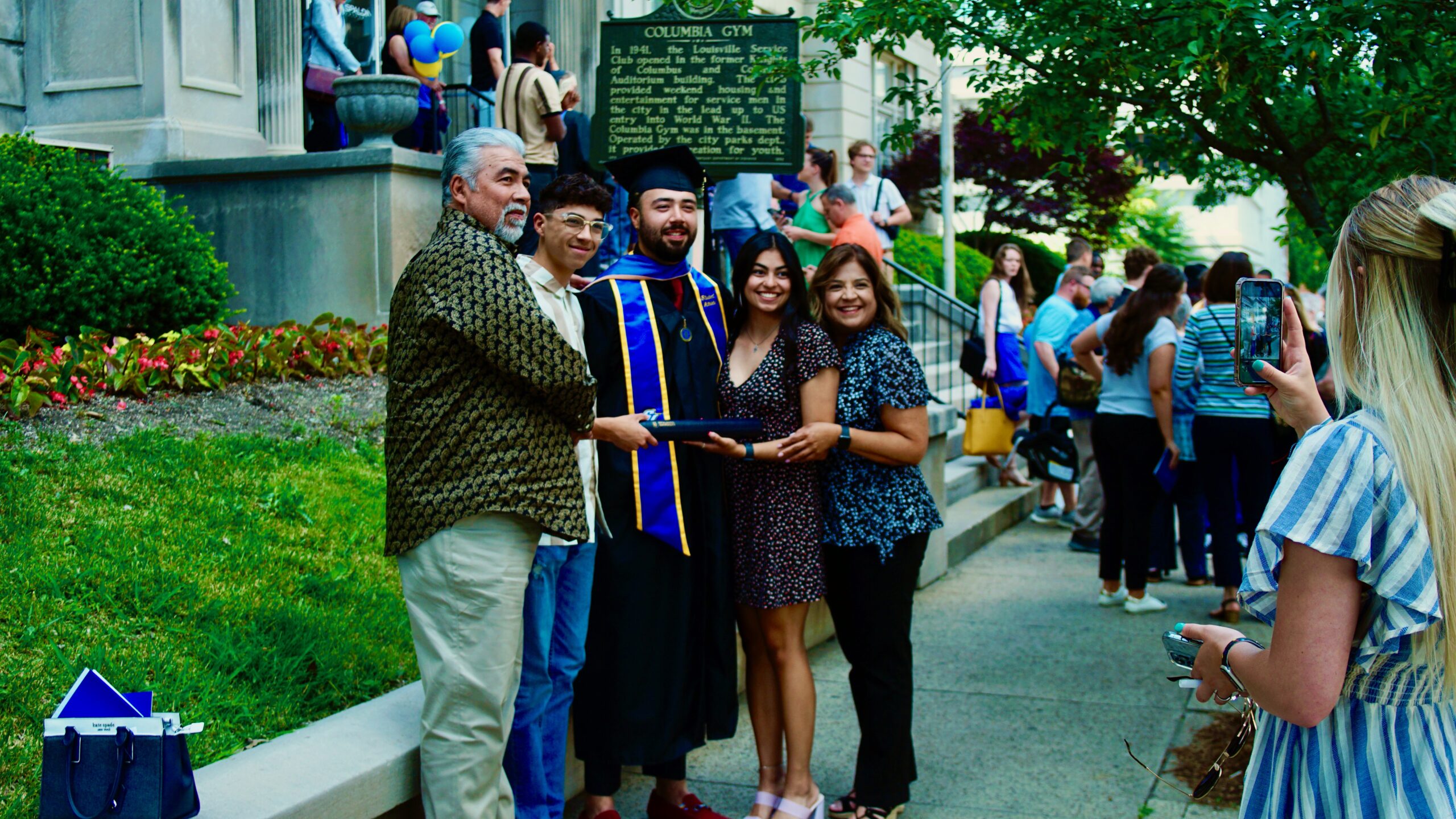 Spalding master's graduate in cap and gown posing outside Columbia Gym with family