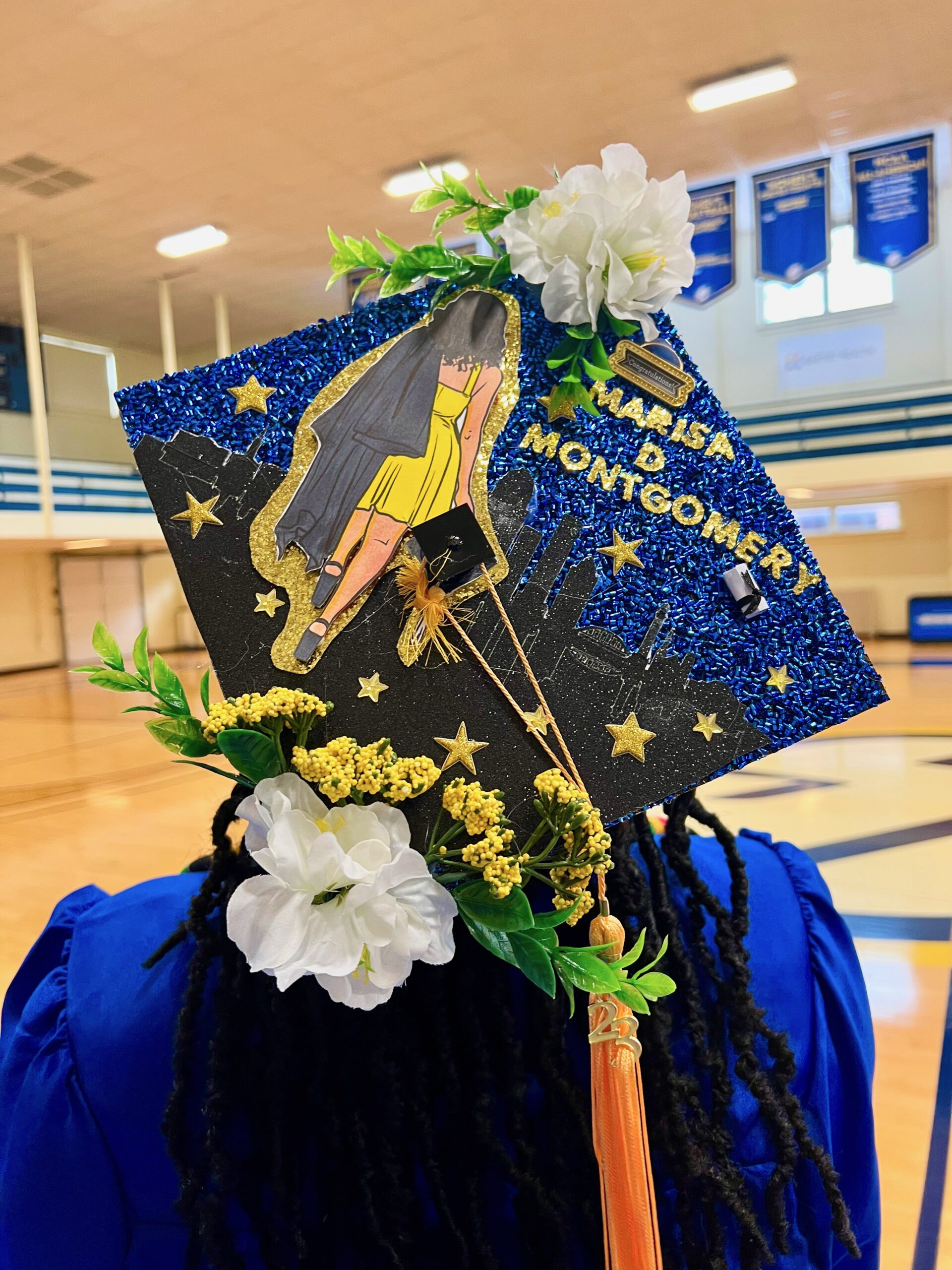 Decorated mortarboard reading "Marisa D Montgromery" wiith picture of the back of a woman in yellow dress walking towards the silhouette of the Louisville skyline while she holds a cap and gown.