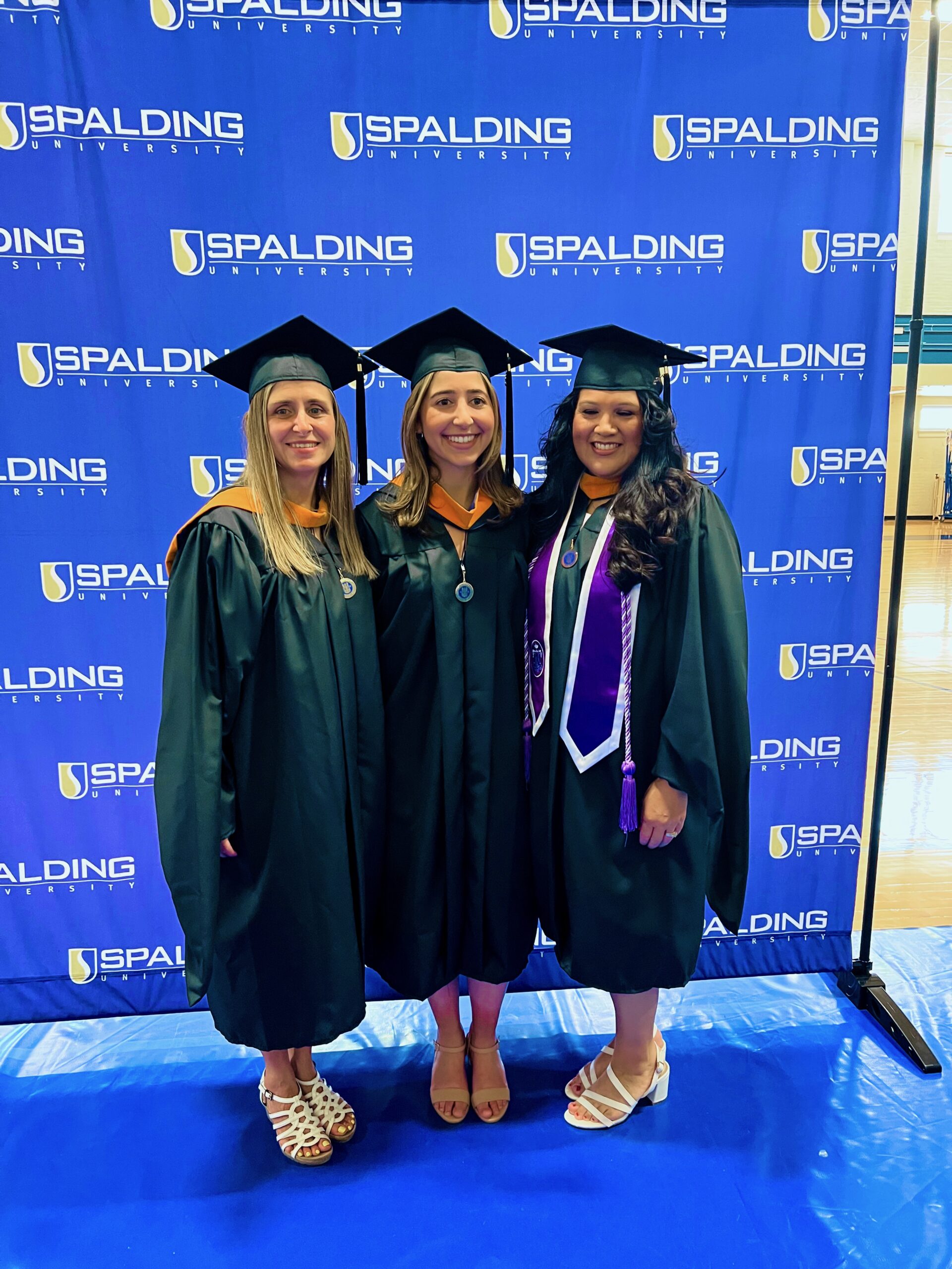 Three Spalding master's graduates posing in front of Spalding backdrop in Columbia Gym before ceremony