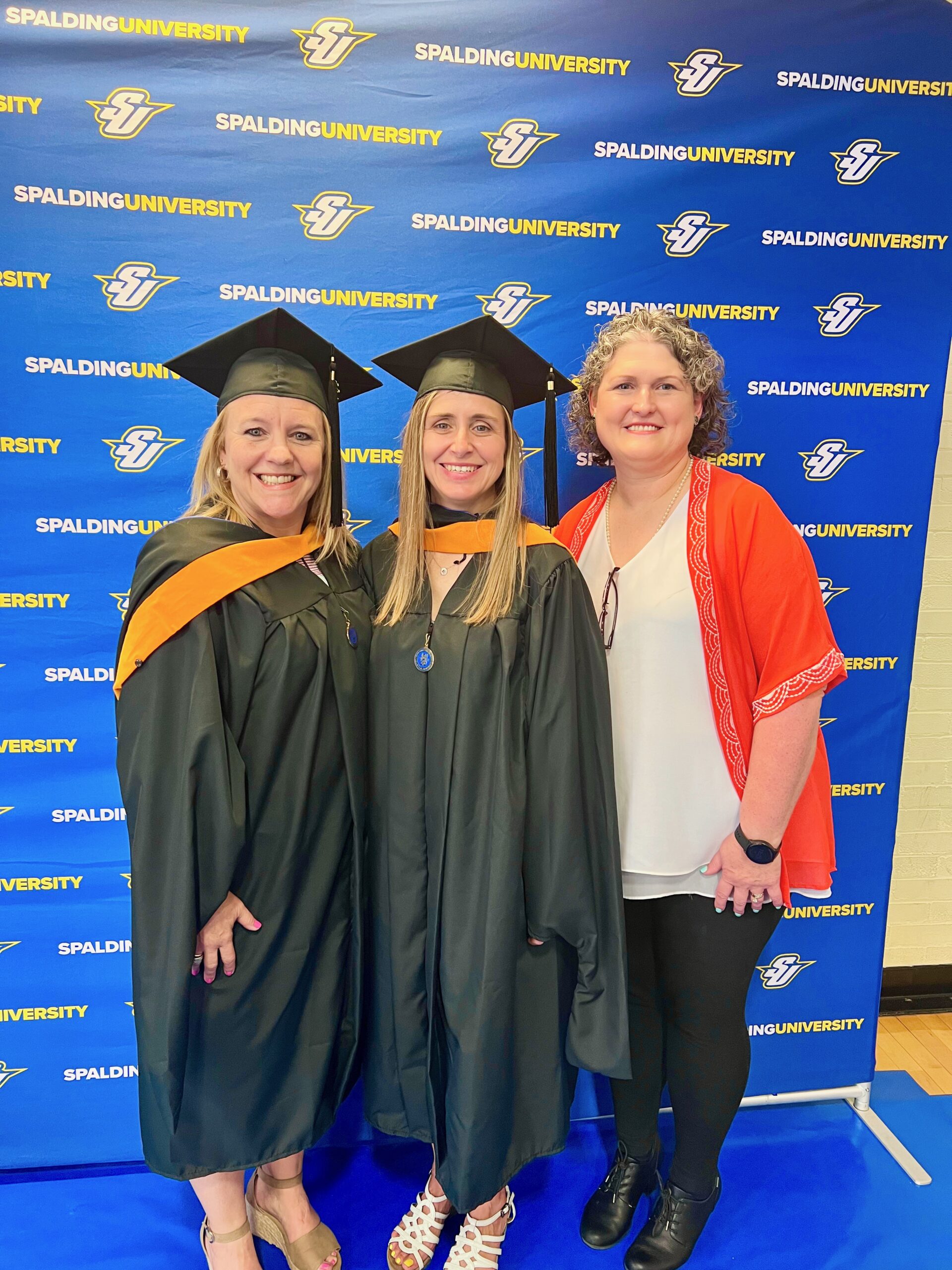 Two Spalding master's graduates posing with mom in front of Spalding Athletics backdrop