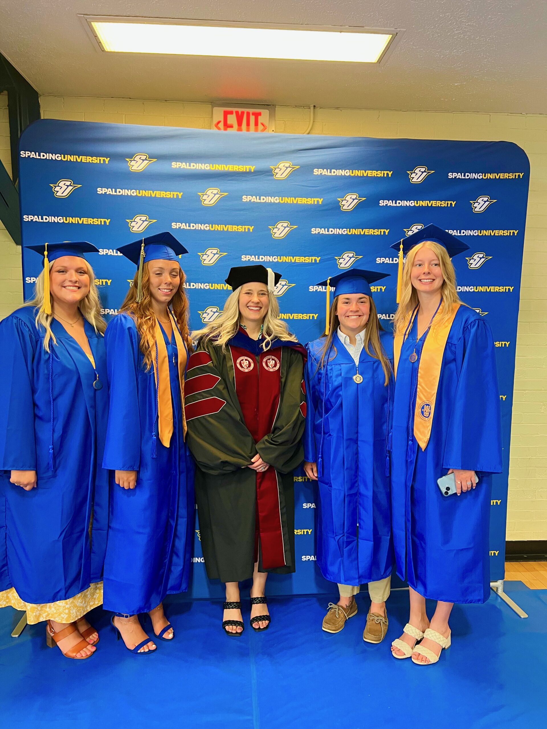 Spalding professor posing with group of bachelor's graduates in front of Spalding Athletics backdrop