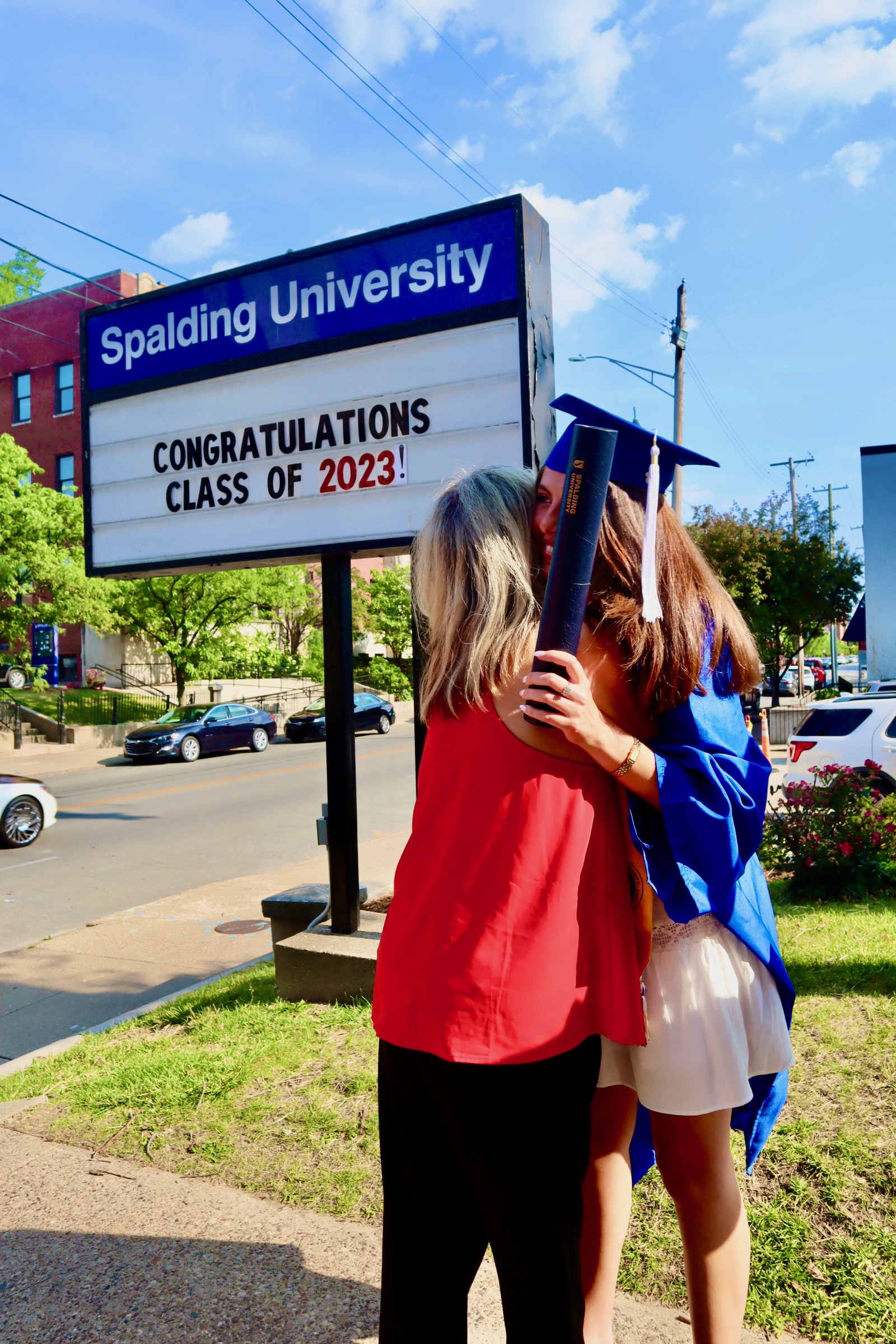 Spalding bachelor's graduate holding degree tube hugging mom in front of sign reading "Congratulations Class of 2023"
