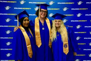 Three Spalding bachelor's graduates posing for photo in front of Spalding Athletics backdrop