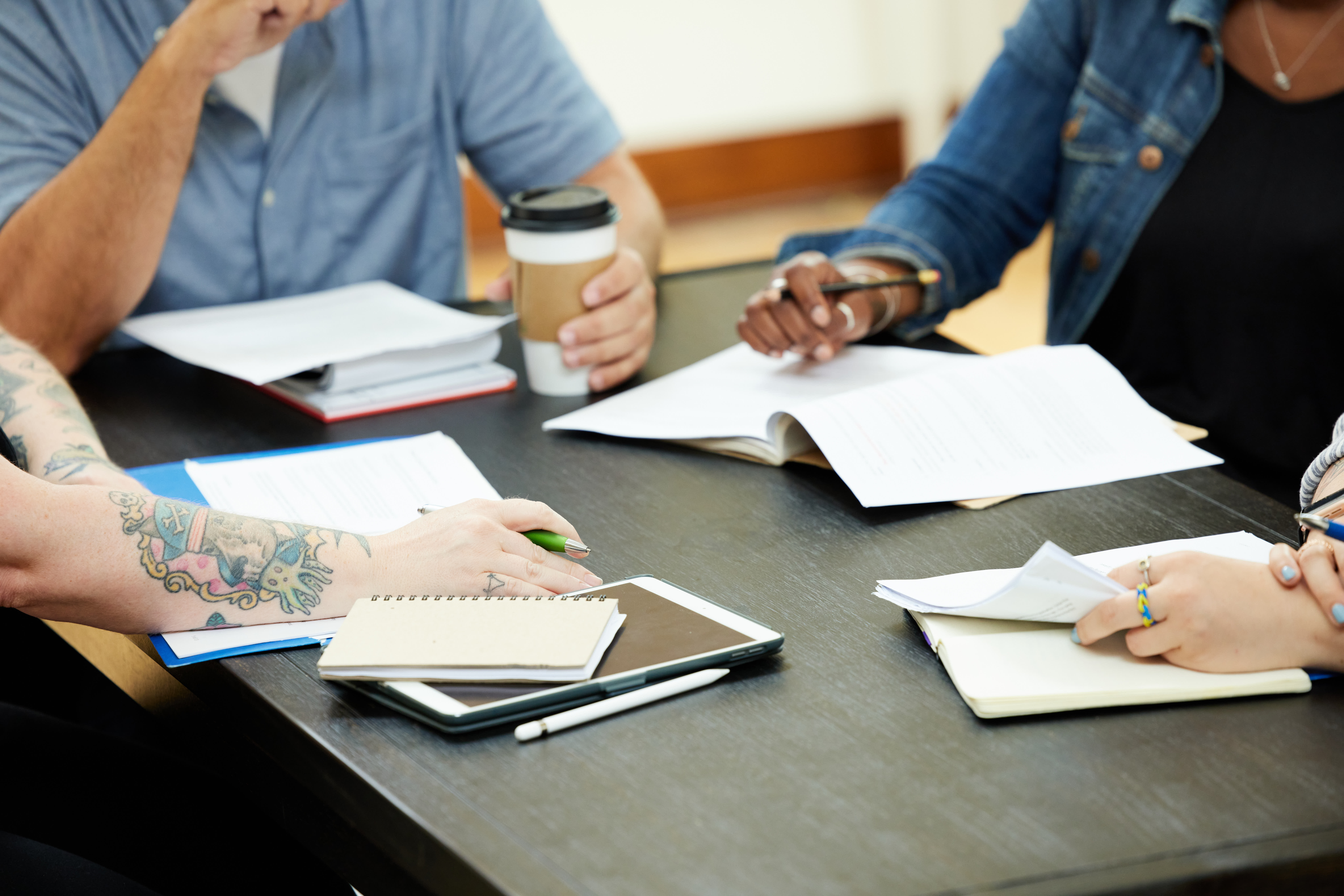 Writers around a table, no faces showing. Focus is on the hands of each writer. Two are holding writing implements with their hands resting on notepads. A third is holding a cup of coffee.