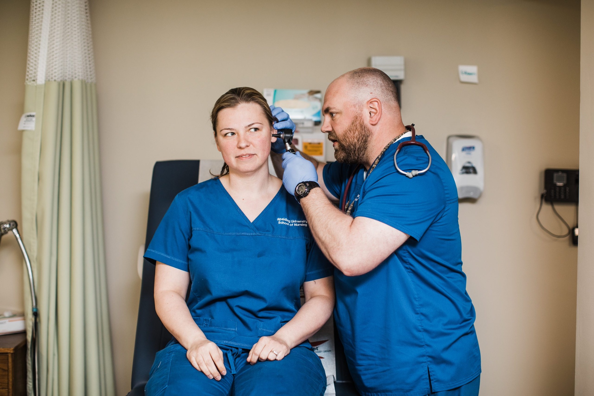 Two Spalding University nursing students in blue scrubs practicing an ear exam