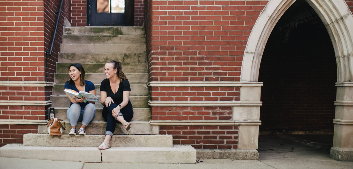 two students sitting on steps in front of brick building with arch