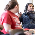 Two male students have a conversation in class while sitting with open books and computers
