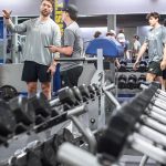 Male trainer in a long sleeve gray shirt provides feedback to two male students in the weight room
