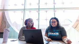 Two black female students visit in front of a laptop computer
