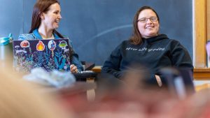 Two female students laugh during a class discussion; laptops are on a table in front of them