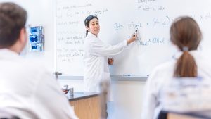Female professor in white lab coat at whiteboard in biology classroom