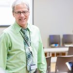 Caucasian male with white hair and a dress shirt and tie talks to two students in a classroom