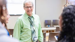 Caucasian male with white hair and a dress shirt and tie talks to two students in a classroom
