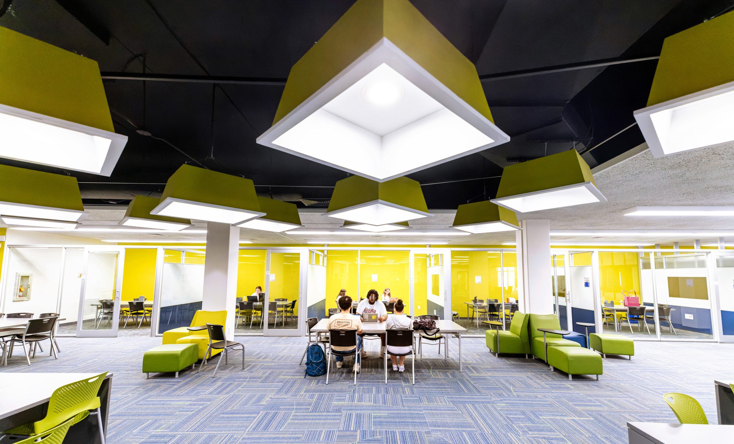 Interior shot of a large, modern common study area with yellow walls and furniture; three students are studying together at a table.