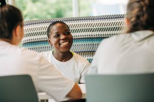 Black female student smiles at two other female students sitting across a table
