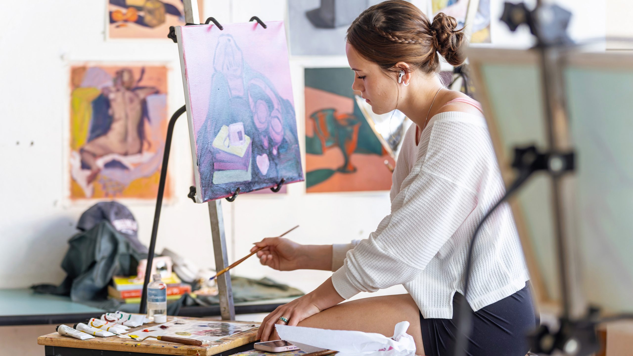 Female student in a white shirt paints at an easel in a bright studio environment