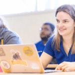 Female student with a blue Spalding polo shirt sits with her laptop in a bright classroom setting