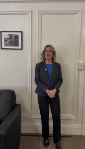 Middle age woman with blonde hair in a gray suit stands in an office talking directly to camera