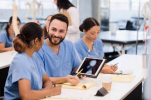Three students in light blue scrubs review x-ray images on a tablet sitting in a classroom